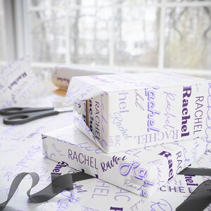 Puede incluir: Papel de regalo blanco con el nombre "Rachel" impreso en letra cursiva morada. El nombre se repite varias veces en el papel de regalo.