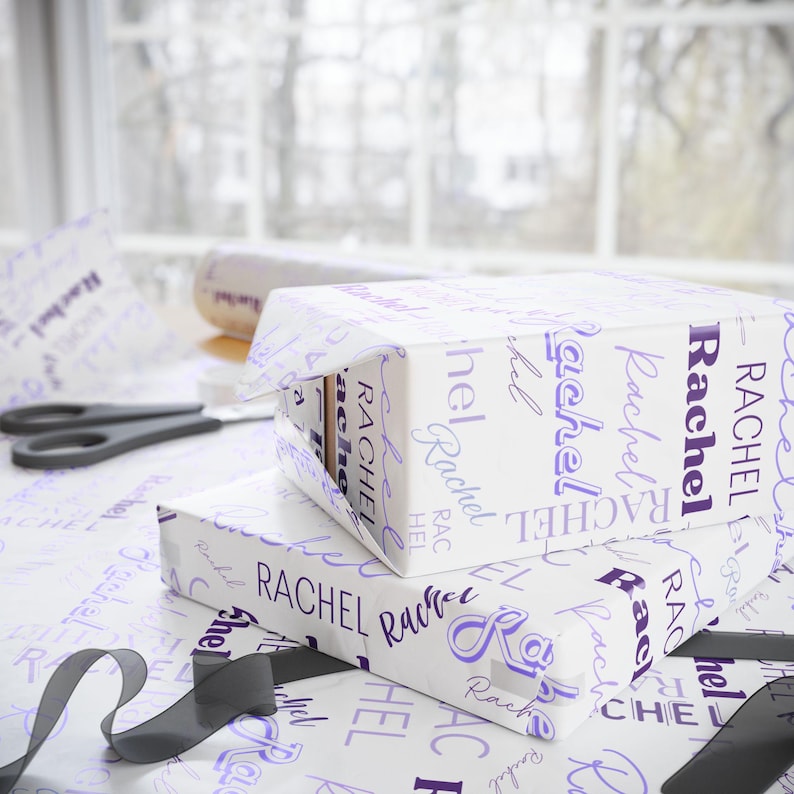 Puede incluir: Papel de regalo blanco con el nombre "Rachel" impreso en letra cursiva morada. El nombre se repite varias veces en el papel de regalo.