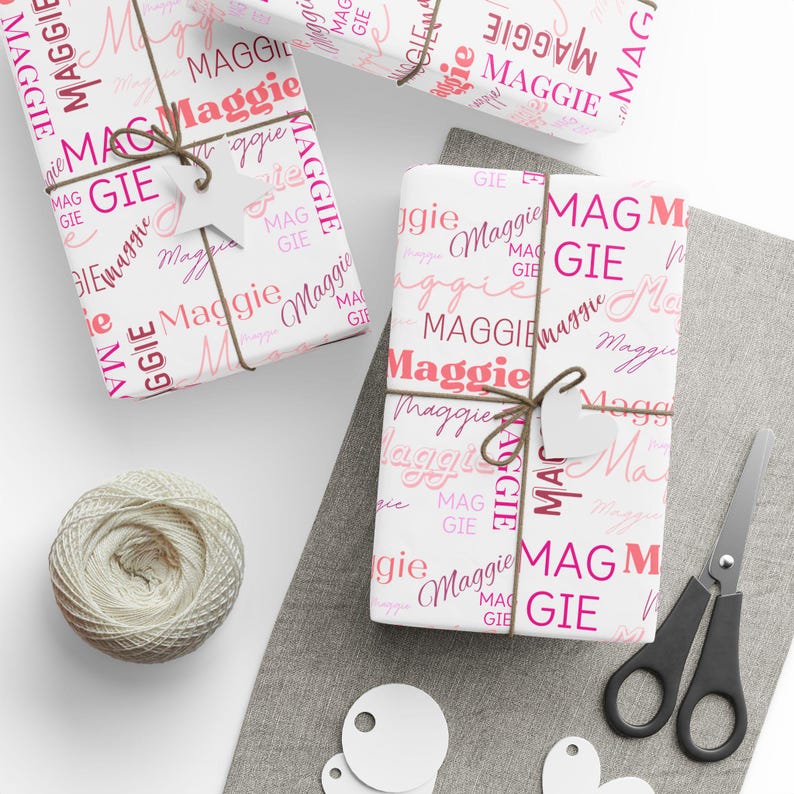 Puede incluir: Papel de regalo blanco con un patr&oacute;n rosa y rojo del nombre "Maggie" repetido en todo. El papel est&aacute; envuelto alrededor de una caja de regalo y atado con una cuerda.