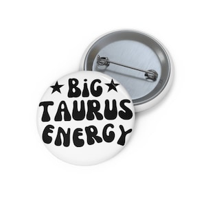 Puede incluir: Botón blanco con texto negro que dice "*BIG* TAURUS ENERGY" en una fuente retro.
