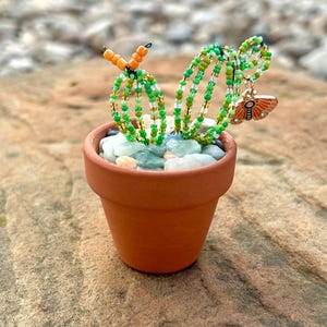 Könnte beinhalten: Ein kleiner Terrakotta-Topf mit einer Perlen-Kaktuspflanze und einem Schmetterlings-Anhänger. Der Kaktus besteht aus grünen und goldenen Perlen und der Schmetterling ist orange und braun.