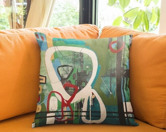 Puede incluir: Una almohada de tiro colorida con formas geométricas abstractas en tonos de verde, azul, rojo y blanco sobre un fondo blanco. La almohada está sentada en un sofá naranja brillante.