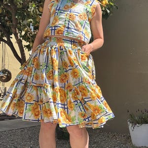 May include: A two-piece outfit featuring a sleeveless top and a tiered skirt. The set is patterned with yellow sunflowers on a white background with blue and white plaid lines. The outfit is paired with platform sandals.