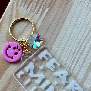 May include: A clear acrylic keychain with the text "FEAR MIND KILL FEAR" in white lettering. The keychain has a gold ring and a pink smiley face charm.