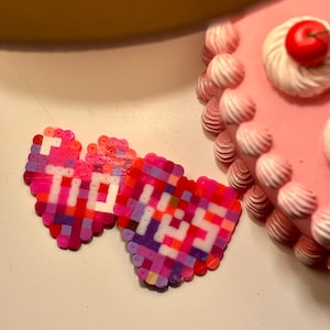 May include: Two pink heart-shaped pixel art pieces with the words "TO" and "KISS" spelled out in white beads.