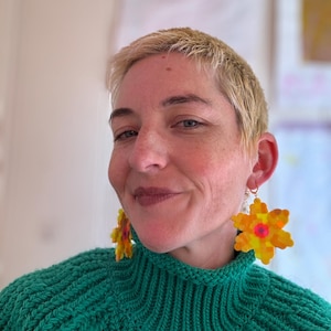 May include: A person with short blonde hair wearing a green knitted sweater and large yellow flower earrings.
