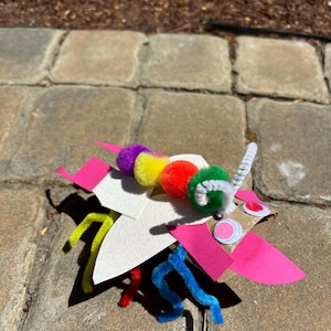 May include: A colorful paper craft bug made from a cardboard tube, pipe cleaners, pom-poms, and construction paper. The bug has pink wings, white antennae, and a rainbow of pom-poms for its body.