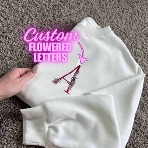 May include: White sweatshirt with a custom embroidered floral letter A. The sweatshirt is a light white color and the letter is a dark pink with small white flowers.