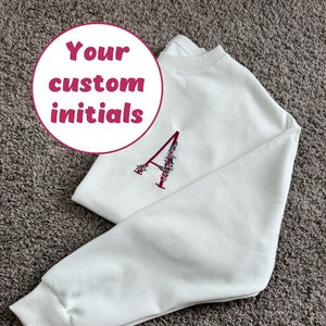 Może przedstawiać: Biała bluza z haftowaną literą A ozdobioną kwiatami. Bluza ma okrągły dekolt i długie rękawy. Tekst "Your custom initials" jest napisany na białym kółku.