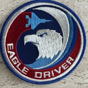 May include: A blue, red and white embroidered patch featuring a white eagle head and a blue jet plane. The text "EAGLE DRIVER" is embroidered in white on a red background.