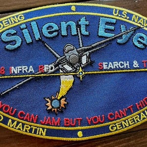 Puede incluir: Parche bordado azul ovalado con el texto "Silent Eye" y la imagen de un avión. El texto adicional incluye "U.S. Navy", "Search & Track" y "You Can Jam But You Can't Hide". El parche también muestra los nombres de Boeing, Lockheed Martin y General Electric.