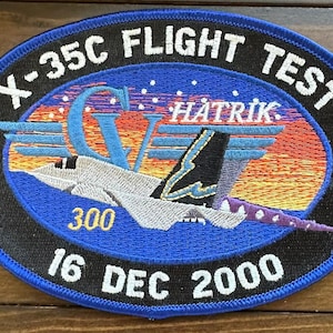 Puede incluir: Un parche bordado azul y negro con el texto "X-35C Flight Test" y "16 Dec 2000". El parche presenta una aeronave blanca con una cola azul y morada, y también se puede ver el texto "Hatrik" y "300".