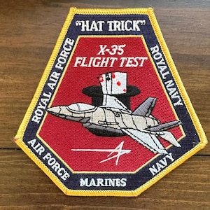 Puede incluir: Un parche bordado en rojo, blanco y azul con un avión de combate volando sobre un sombrero de copa con cartas de juego. El parche tiene el texto "HAT TRICK" y "X-35 FLIGHT TEST" en blanco. El texto "ROYAL AIR FORCE", "AIR FORCE", "MARINES", "ROYAL NAVY" y "NAVY" están en amarillo.