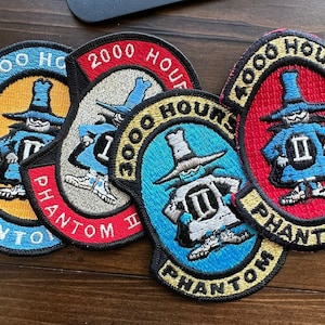 Puede incluir: Cuatro parches bordados con el texto "1000 Hours Phantom II" en azul, rojo y amarillo. Cada parche presenta un personaje de dibujos animados con un traje azul y blanco y un sombrero.