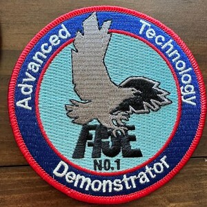 May include: A circular patch with a blue border and red trim. The patch features the words "Advanced Technology" and "Demonstrator" in white text. A gray eagle graphic is centered above the word "FIVE" and "NO.1". The background is light blue.