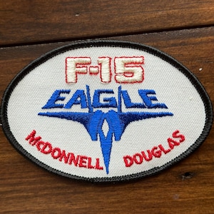 May include: A white, round patch with a black border. The patch features a blue and white F-15 Eagle silhouette with the text "F-15 EAGLE" in red and white lettering. The text "MCDONNELL DOUGLAS" is written in red lettering around the bottom of the patch.