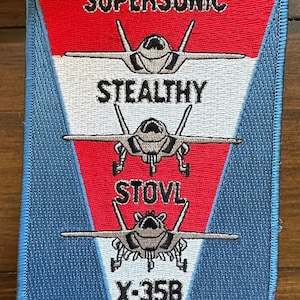 Może przedstawiać: Naszywka haftowana w kolorze niebieskim, białym i czerwonym z samolotem myśliwskim w środku. Na naszywce widoczny jest tekst "SUPERSONIC", "STEALTHY", "STOVL" i "X-35B".