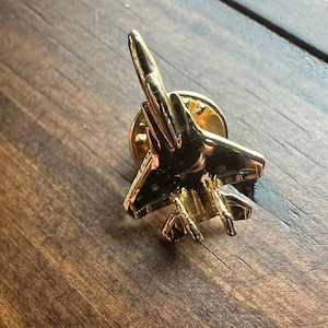 May include: A gold-coloured metal pin depicting a fighter jet. The pin features detailed wings, fuselage, and tail fins. It is set against a dark wooden surface, with the pin's clasp visible.