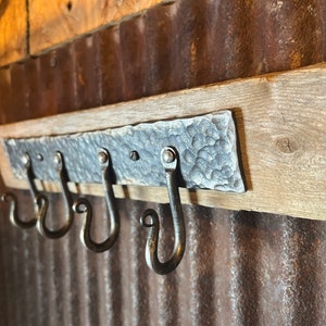 May include: Rustic wooden key holder with three metal hooks. The hooks are attached to a hammered metal plate. The holder is mounted on a corrugated metal wall.