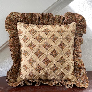 May include: A square throw pillow featuring a beige fabric overlay with a circular lattice pattern revealing a small-scale floral print in shades of orange, green, and brown underneath. The pillow is finished with a wide, ruffled fabric border.