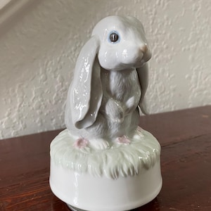 May include: A porcelain music box featuring a light gray lop-eared rabbit with long ears and dark eyes. The rabbit sits on a textured, pale green base that resembles grass, which is mounted on a smooth, cylindrical white ceramic pedestal base.