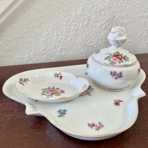 May include: A white porcelain vanity set featuring a large, irregularly shaped tray, a small oval dish, and a lidded jar. Each piece is decorated with delicate, colourful floral patterns and finished with a thin gold trim along the edges.