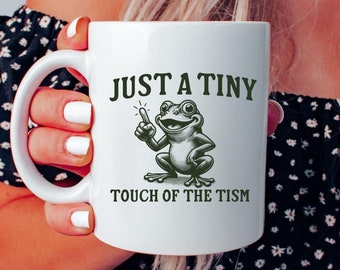 Può includere: Una tazza in ceramica bianca con un'illustrazione di rana verde e il testo "Just a Tiny Touch of the Tism".