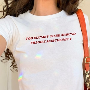 Peut inclure: Un t-shirt blanc court avec le texte "Too Clumsy to be Around Fragile Masculinity" en lettres rouges. Le t-shirt est porté par une personne avec un sac en cuir marron avec une bandoulière orange.