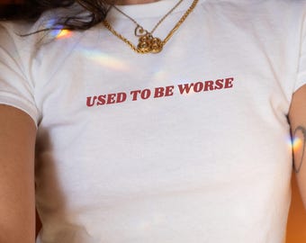 May include: White t-shirt with the phrase "USED TO BE WORSE" printed in red. The shirt is a crew neck style. A gold necklace is worn around the neck.