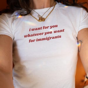 Pode incluir: Camiseta branca com o texto "i want for you whatever you want for immigrants" em vermelho. A camiseta é de manga curta e tem gola redonda. Um colar de ouro é usado no pescoço.