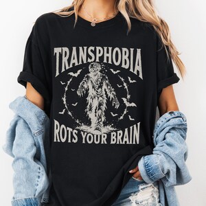 以下が含まれることがあります： 白のグラフィックプリントが施された黒の半袖Tシャツ。中央に描かれたクリーチャーの周りにコウモリが配置され、その上下に「TRANSPHOBIA ROTS YOUR BRAIN」という文字がかすれたアーチ状のフォントでプリントされています。