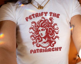 Pode incluir: Camiseta branca com um gráfico vermelho de Medusa e o texto "PETRIFY THE PATRIARCHY". A ilustração de Medusa apresenta cobras como cabelo. A camiseta tem gola redonda e mangas curtas.