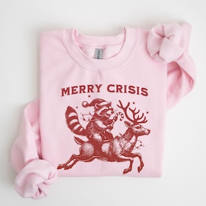 May include: A light pink sweatshirt with a red graphic. The graphic features a raccoon wearing a Santa hat and riding a reindeer, holding a candy cane. The text "MERRY CRISIS" is above the graphic.