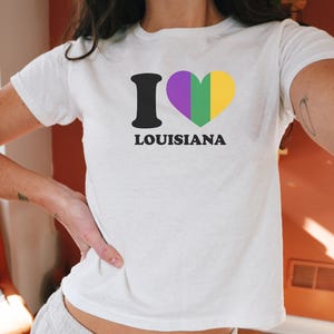Puede incluir: Camiseta blanca con el texto "I Louisiana" en negro, con un símbolo de corazón en morado, verde y amarillo. La camiseta es de manga corta y está hecha de un material suave.