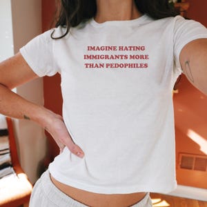May include: White t-shirt with the text "IMAGINE HATING IMMIGRANTS MORE THAN PEDOPHILES" in red letters. The shirt is short-sleeved and appears to be made of cotton or a similar fabric.