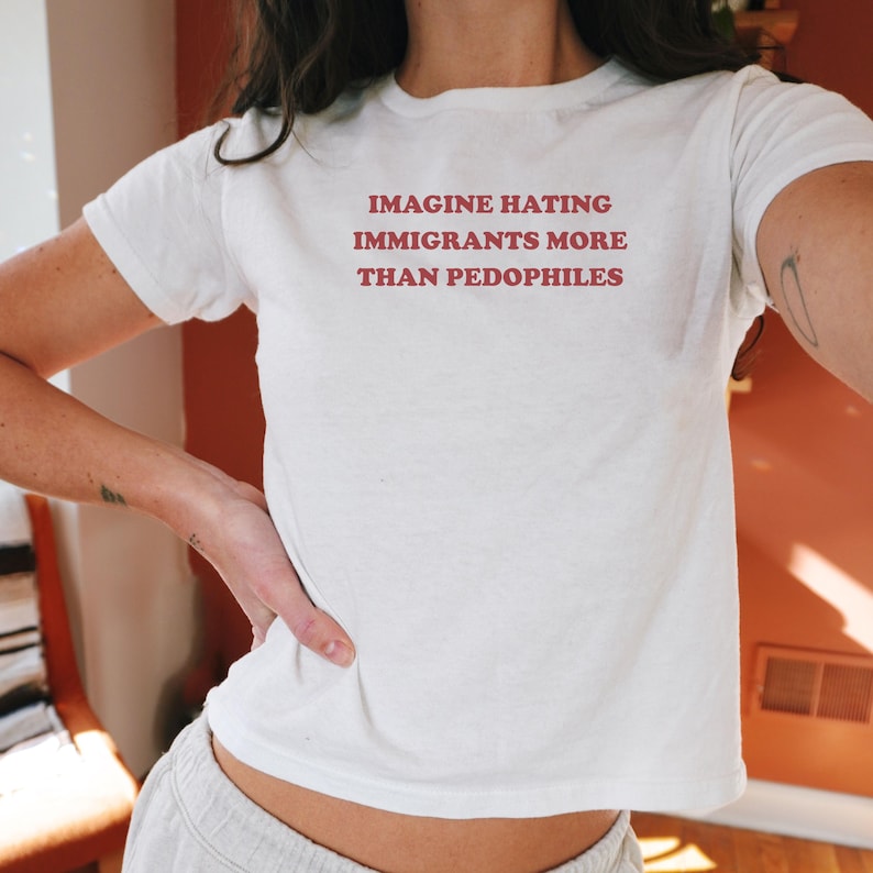 May include: White t-shirt with the text "IMAGINE HATING IMMIGRANTS MORE THAN PEDOPHILES" in red letters. The shirt is short-sleeved and appears to be made of cotton or a similar fabric.
