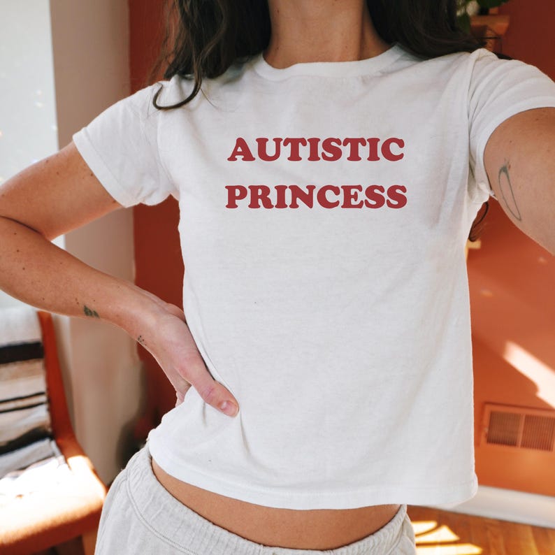 以下が含まれることがあります： 赤い文字で「AUTISTIC PRINCESS」と書かれた白いTシャツです。