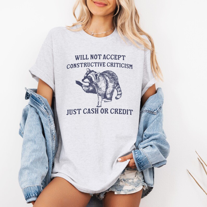 May include: A gray t-shirt with a raccoon graphic and the text "Will not accept constructive criticism. Just cash or credit."