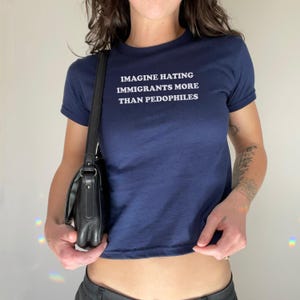 May include: Navy blue crop top with the text "IMAGINE HATING IMMIGRANTS MORE THAN PEDOPHILES" in white. The shirt has short sleeves and a crew neck. A black shoulder bag is visible.