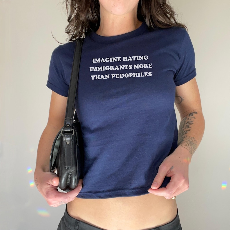 May include: Navy blue crop top with the text "IMAGINE HATING IMMIGRANTS MORE THAN PEDOPHILES" in white. The shirt has short sleeves and a crew neck. A black shoulder bag is visible.