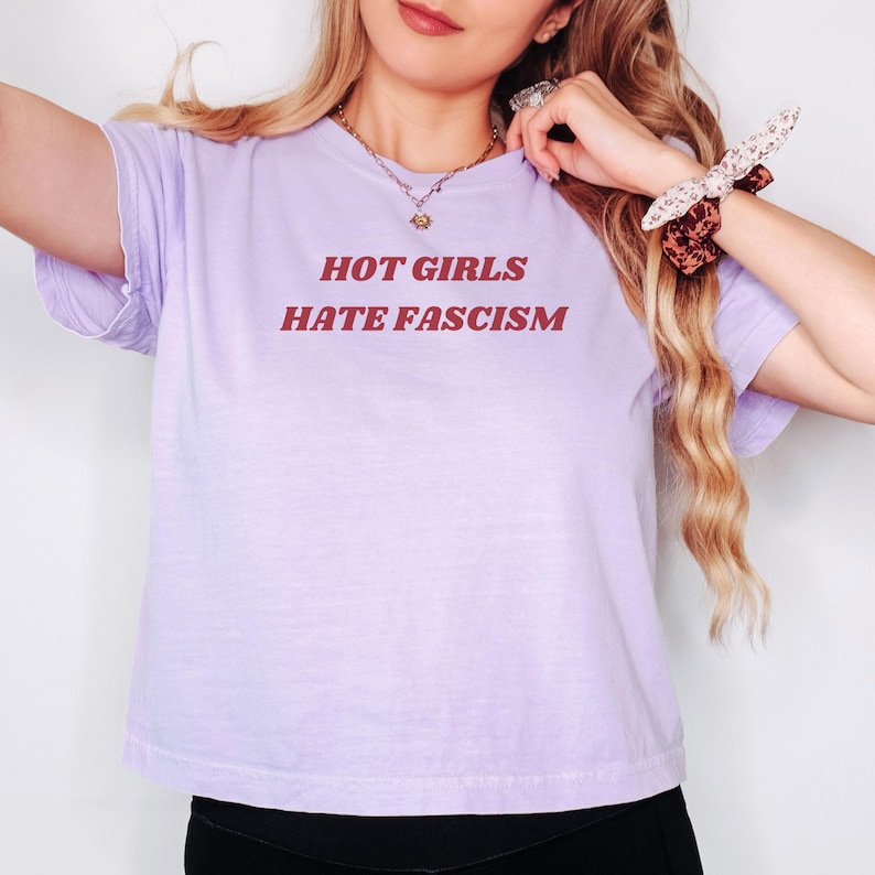 Puede incluir: Una camiseta de color lila claro con el texto rojo "HOT GIRLS HATE FASCISM". La camiseta tiene mangas cortas. La persona lleva un collar y una goma para el pelo estampada. La camiseta es de color liso.