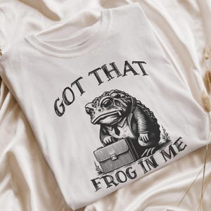 Puede incluir: Camiseta blanca con una ilustraci&oacute;n en blanco y negro de una rana con traje y sosteniendo un malet&iacute;n. El texto "Got that frog in me" est&aacute; impreso en la camiseta.