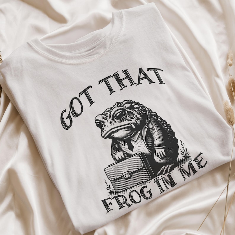 Puede incluir: Camiseta blanca con una ilustraci&oacute;n en blanco y negro de una rana con traje y sosteniendo un malet&iacute;n. El texto "Got that frog in me" est&aacute; impreso en la camiseta.