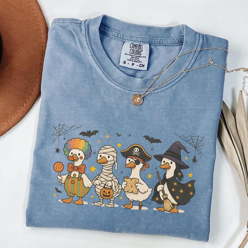 May include: A light blue Comfort Colors t-shirt featuring a Halloween-themed graphic of ducks in costumes. The ducks are dressed as a clown, mummy, pirate, and witch. A gold necklace and a brown hat are also visible.