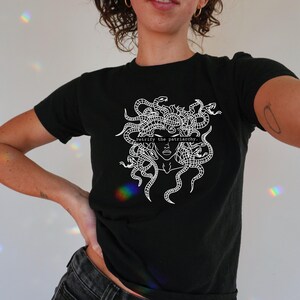 Puede incluir: Camiseta negra con un gr&aacute;fico blanco de la cabeza de Medusa con serpientes como cabello. El texto "Petrify the patriarchy." est&aacute; impreso debajo de la imagen.