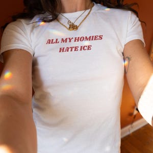 May include: White t-shirt with the text "ALL MY HOMIES HATE ICE" in red letters. The shirt is short-sleeved and appears to be made of cotton. A gold necklace is worn around the neck.