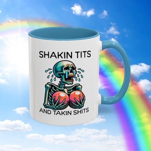 May include: White ceramic mug with a blue interior and handle. The mug features a cartoon skeleton design with the text "SHAKIN TITS AND TAKIN SHITS". The skeleton is crying and holding two pink breasts.