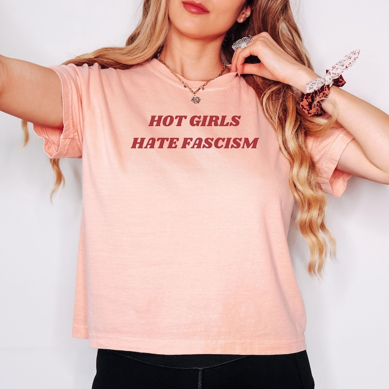 Puede incluir: Una camiseta rosa claro con el texto "HOT GIRLS HATE FASCISM" en fuente rojo oscuro. La camiseta es de cuello redondo y parece estar hecha de un material suave. La est&eacute;tica general es casual y c&oacute;moda.