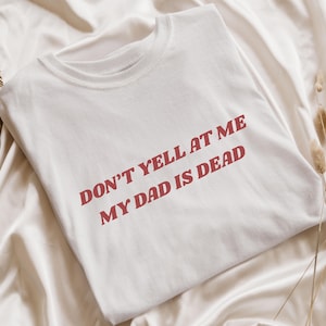 Op de afbeelding: Wit T-shirt met de tekst "DON'T YELL AT ME MY DAD IS DEAD" in rode letters. Het shirt is opgevouwen op een witte satijnen ondergrond, met gedroogde planten en een waaier van palmbladeren op de achtergrond.