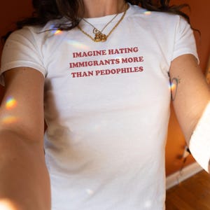 Op de afbeelding: Wit T-shirt met de rode tekst "IMAGINE HATING IMMIGRANTS MORE THAN PEDOPHILES". Het shirt heeft een ronde hals. Een gouden ketting is zichtbaar.